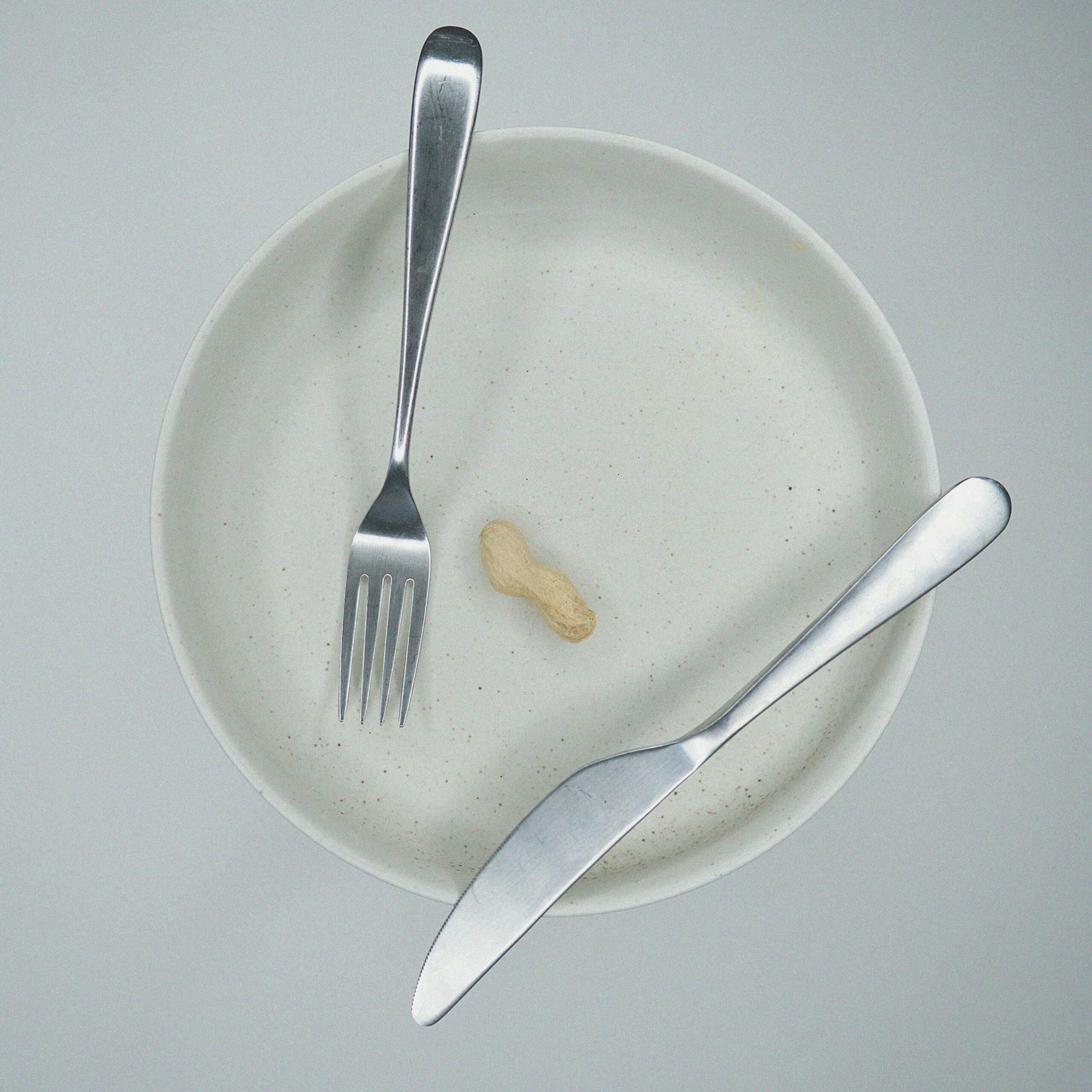 Top view of a white plate with a single peanut and silver utensils on a light background.